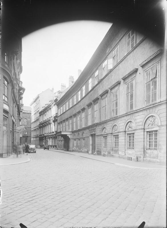 The black-and-white photo captures a historic Vienna street, where the OSCE Secretariat is located now. It features cobblestone roads, elegant multi-story buildings, and a façade with arched windows and decorative reliefs. A shop sign is visible, along with vintage cars and blurred pedestrians. The curved street adds depth, and the dark vignette suggests an old-fashioned camera.